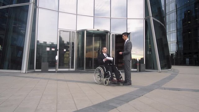 Male adult colleagues in suits talking outdoors, one is on wheelchair, business communication.