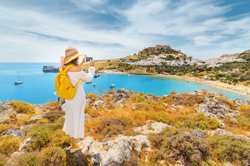 Young woman traveler taking photo of her smartphone scenic landscape of a famous tourist attraction on Rhodes Island - Lindos town - obrazy, fototapety, plakaty