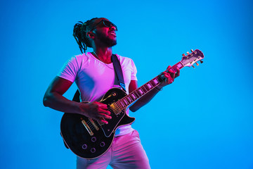 Young african-american musician playing the guitar like a rockstar on blue studio background in neon light. Concept of music, hobby. Joyful attractive guy improvising. Retro colorful portrait. - obrazy, fototapety, plakaty