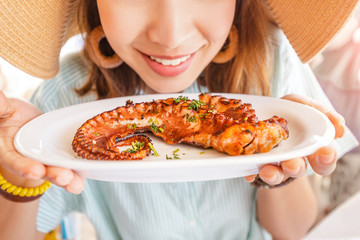 Happy asian woman in hat eating delicious grilled octopus in seafood restaurant. Delicacy and...