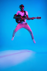 Fototapeta na wymiar Young african-american musician playing the guitar in jump like a rockstar on blue studio background in neon light. Concept of music, hobby. Joyful attractive guy improvising. Retro colorful portrait.