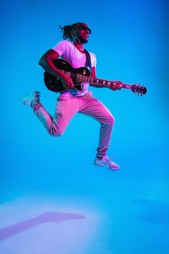 Young African-american Musician Playing The Guitar In Jump Like A Rockstar On Blue Studio Background In Neon Light. Concept Of Music, Hobby. Joyful Attractive Guy Improvising. Retro Colorful Portrait.