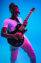 Young african-american musician playing the guitar like a rockstar on blue studio background in...