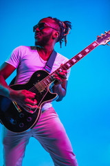 Fototapeta premium Young african-american musician playing the guitar like a rockstar on blue studio background in neon light. Concept of music, hobby. Joyful attractive guy improvising. Retro colorful portrait.