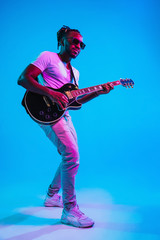 Young african-american musician playing the guitar like a rockstar on blue studio background in...