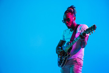 Young african-american musician playing the guitar like a rockstar on blue studio background in...