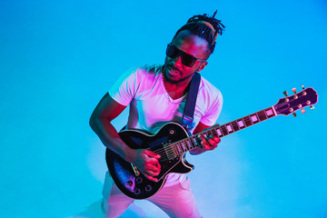 Young african-american musician playing the guitar like a rockstar on blue studio background in neon light. Concept of music, hobby. Joyful attractive guy improvising. Retro colorful portrait.