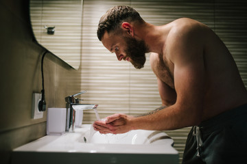 Young bearded hipster man washing his face with water in the bathroom while looking himself in the...