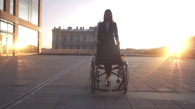 Asian buisness woman rolling Invalid wheelchair near modern building at sunset. Sunny teal and orange steadicam shot.