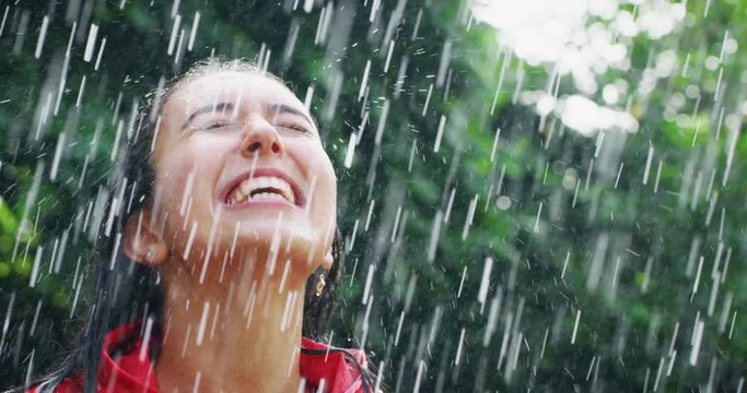 Slow motion of young carefree beaoutiful woman wearing protection cape is feeling free under the rain on a background of green trees.