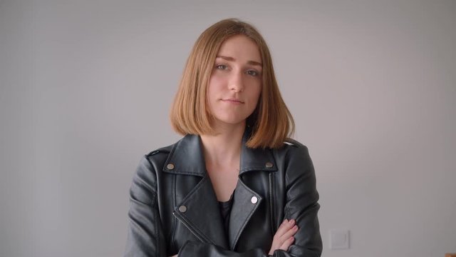 Closeup portrait of young cute caucasian girl in a leather jacket having her arms crossed looking at camera indoors in the empty room