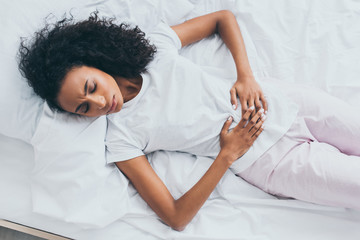 beautiful african american woman suffering from abdominal pain while lying on white bedding