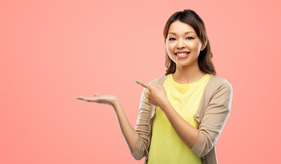 people and advertisement concept - happy asian young woman holding something imaginary on empty hand over pink living coral background