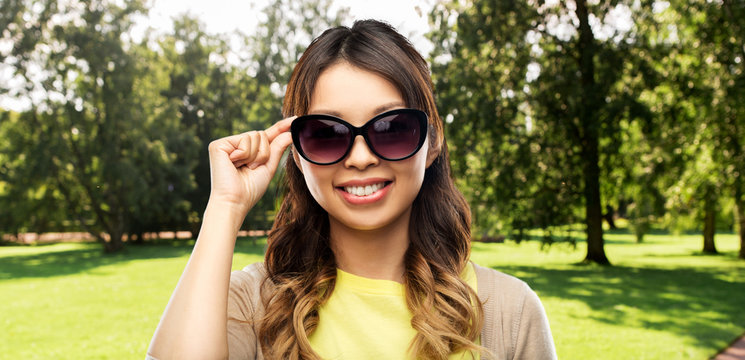 fashion, style and eyewear concept - happy smiling young asian woman in sunglasses over summer park background