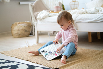 A little two-year-old girl sits on the bedroom floor and plays. Toy calendar in the form of a puzzle. Educational games for children. Bright bedroom, black and white carpet near the bed