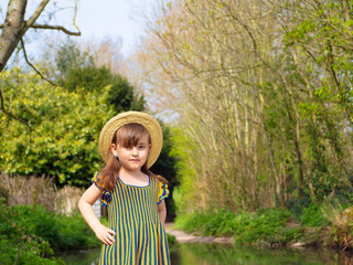 Fototapeta premium Adorable, smiling little girl in rustic dress and straw hat is playing in spring forest