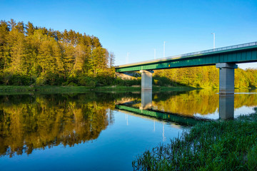Beautiful river with a forest, the reflection of trees in the water, smooth calm surface of the water without waves. View of the bridge over the river.