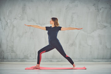 Attractive fit Caucasian brunette in sportswear doing Warrior II yoga pose on mat barefoot.