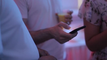 A man in a white T-shirt looks into his phone at a party. A man holds in his hand a smartphone and a glass of orange juice. Light from the phone screen. Dim light in the room