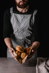 A young man with a beard shows freshly baked buns. Black background.