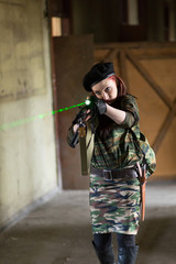 Fototapeta premium A young woman in an abandoned building with a gun. Laser sight on the machine. Aiming from the machine with a laser. The girl in the black beret, in green camouflage.