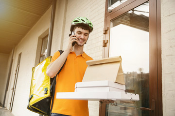 Fototapeta na wymiar Young man in yellow shirt delivering pizza using gadgets to track order at the city's street. Courier using online app for receiving payment and tracking shipping address. Modern technologies.