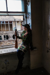 Fototapeta premium A young woman with a rifle in uniform at the window looking at the street. The woman sniper in a green suit and cap.