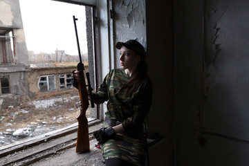 A young woman with a rifle in uniform at the window looking at the street. The woman sniper in a green suit and cap.