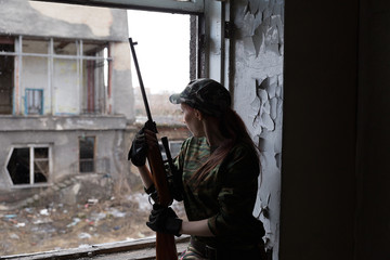 A young woman with a rifle in uniform at the window looking at the street. The woman sniper in a green suit and cap.