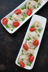 Top view of dumplings with green peas and cherry tomatoes served on two white plates, vertical shot on a dark brown stone background