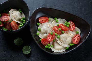 Black bowls with ravioli served with cherry tomatoes and green peas, studio shot on a black stone surface