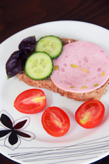 Sandwich of rye bread with delicious sausage, slices of fresh cucumber, tomato and basil leaves on a white plate