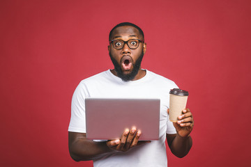 Young surprised african man standing and using laptop computer isolated over red background. Drinking coffee or tea.