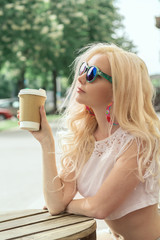 Beautiful blonde girl in the city is drinking coffee. Street photo session. Gray cup with a white lid and a place for the logo