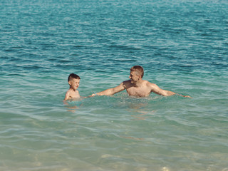 Cute European boy is splashing in the sea with his dad. They are having fun during their holidays on the seacoast.