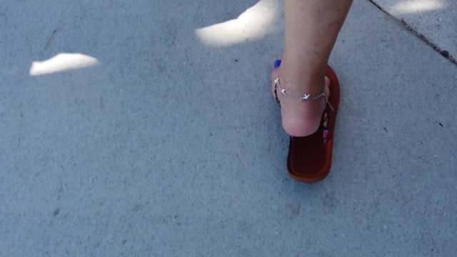 Slow motion footage of a woman's feet in flat sandals with blue nail polish walking on concrete sidewalk