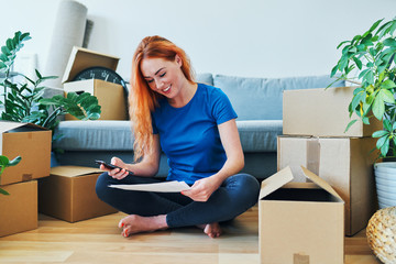 Young woman sitting on floor of new apartment with smartphone and bills