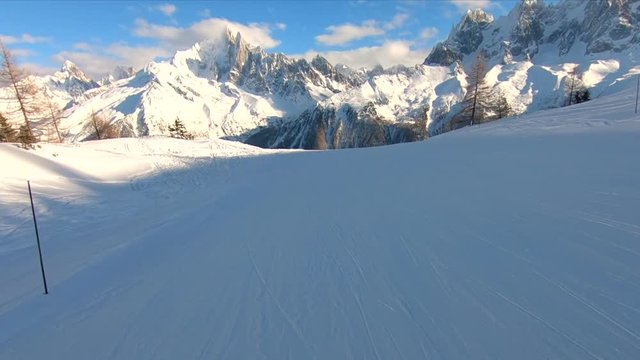 This is a moving shot on a ski slope with mountains in the background and lots of foreground. There is also sunshine blue sky and clouds on top of the mountains.