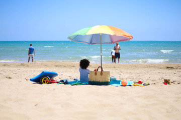 BIMBO DI SPALLE IN SPIAGGIA SOTTO OMBRELLONE COLORATO CON I GIOCHI CHE GUARDA VERSO IL MARE