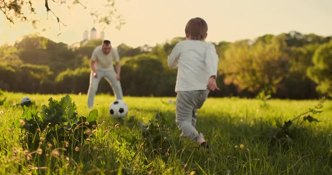 Father standing at the gate playing football with his son at sunset. The father trains the child on the field. Boy strikes the ball.