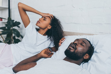 young african american woman suffering from headache while lying near snoring husband