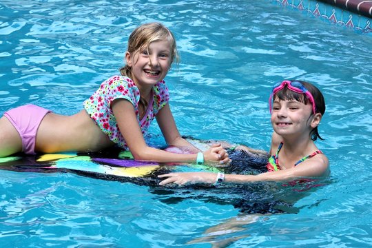 Two girls having fun in the pool with a kickboard smiling at the camera