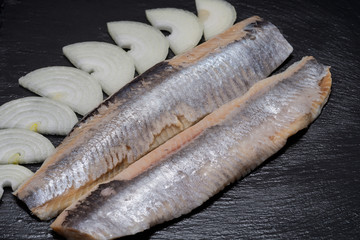 Salted boneless fillets of wild-caught Atlantic Herring and sliced Yellow Onion arranged on natural black stone background. Clupea harengus.
