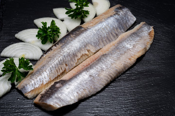 Delicious Salted Herring garnished with Yellow Onion slices and Fresh Parsley. Natural black stone background. Clupea harengus.
