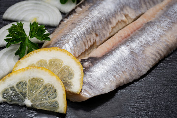 Salted Deboned Herring Fillets garnished with Yellow Onion, Lemon and Fresh Parsley. Natural black stone background. Clupea harengus.