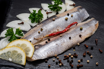 Salted Herring Fillets garnished with Yellow Onion, Lemon, Fresh Parsley and Dried Chili Pepper. Natural black stone background. Clupea harengus.