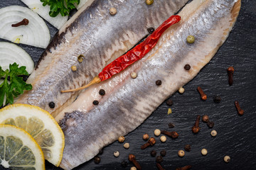 Salted Herring Fillets garnished with Yellow Onion, Lemon, Fresh Parsley and Dried Chili Pepper. Natural black stone background. Clupea harengus.