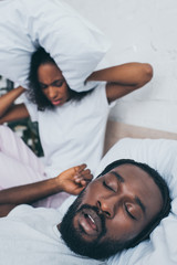 Fototapeta na wymiar selective focus of african american woman covering head with pillow while sitting in bed near snoring husband
