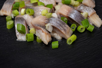 Slices of Salted Boneless wild-caught Atlantic Herring garnished with Chopped Green Onion Scallions. Black natural stone background. Clupea harengus. 