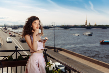 Young beautiful girl with curly hair in a pink dress is standing on the balcony and drinking coffee. Happy woman smiling and looking at the Neva River in St. Petersburg